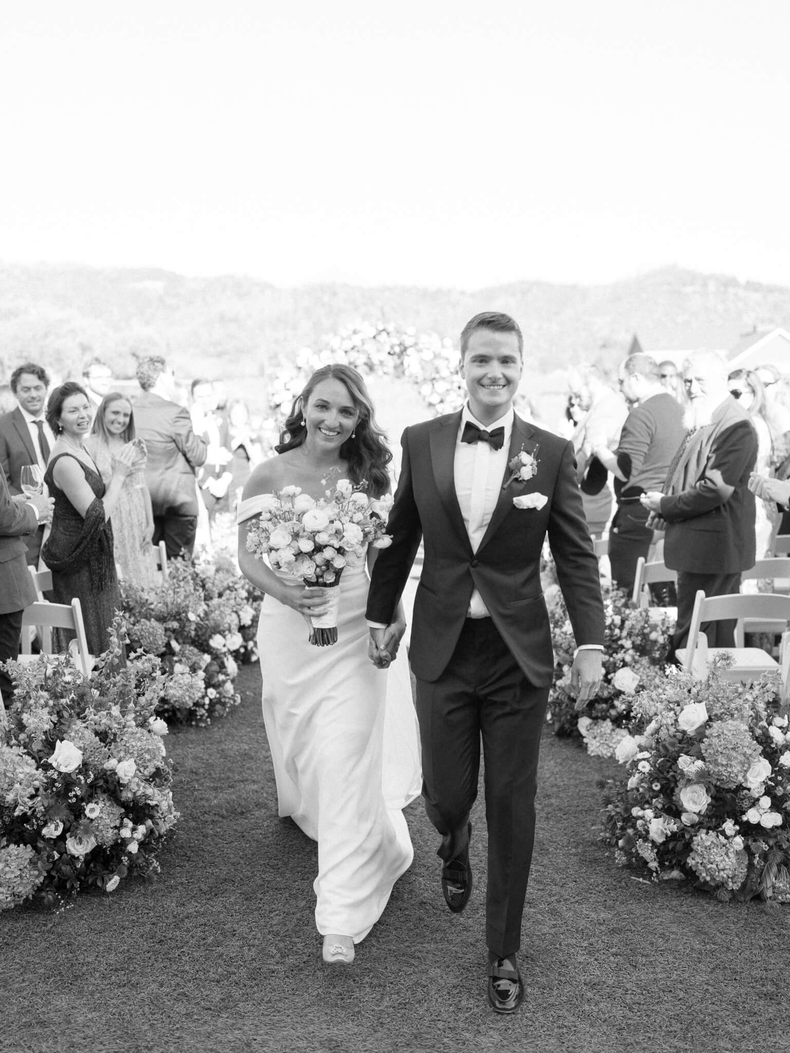 A black and white image of a bride and groom walking away from their ceremony hand in hand with smiling guests looking on.
