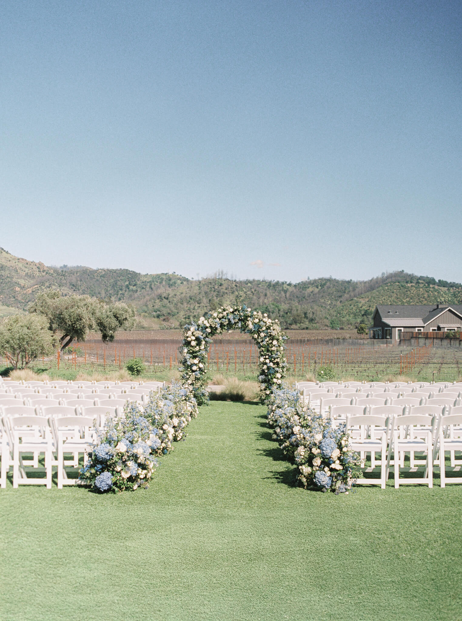 A wedding ceremony set up with white chairs and flowers lining the aisle leading to a floral arch of blue and white flowers.
