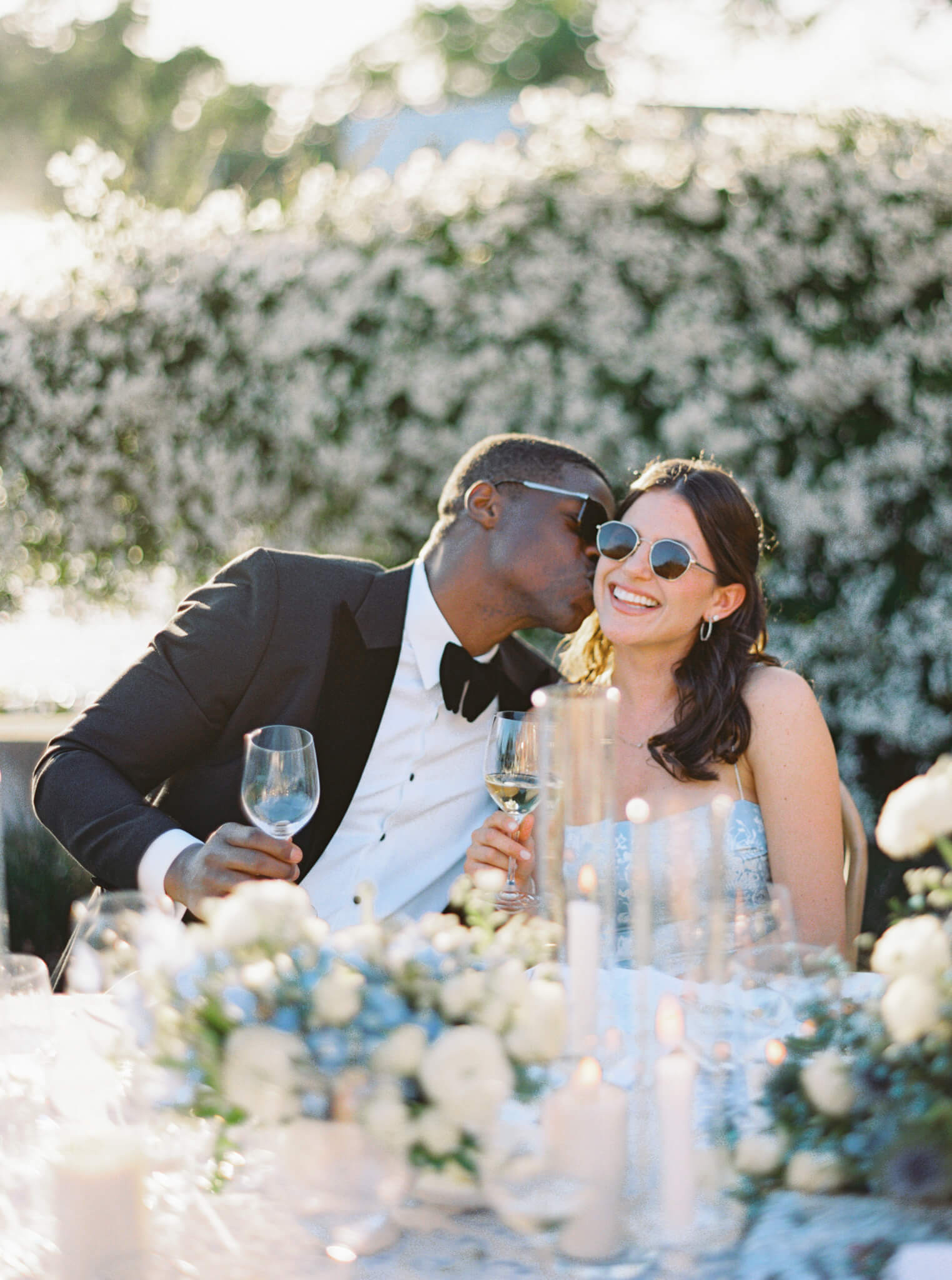 A groomsman kissing a bridesmaid on the cheek at a destination wedding reception surrounded by florals and candles.