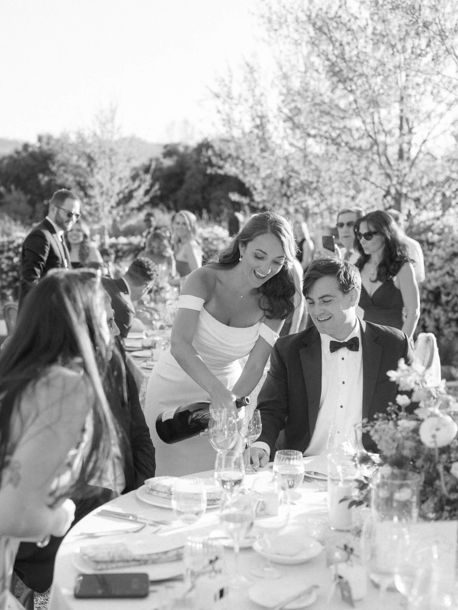 A black and white image of a bride pouring champagne for her guests at her own wedding reception.