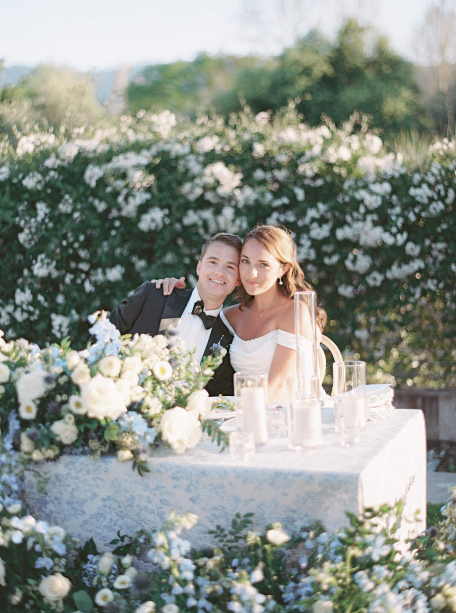A bride and groom leaning into each other at their sweetheart table with lush blue, white and green florals.