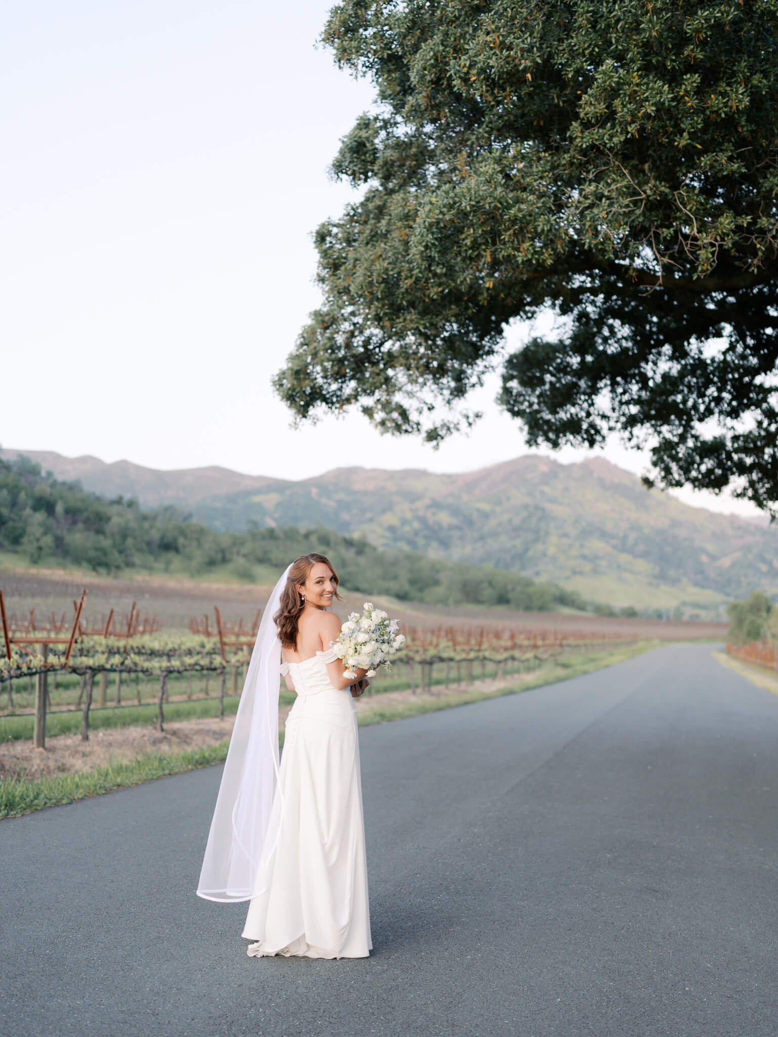 Solo image of a bride on her wedding day facing away from the camera and looking over her shoulder with mountains and vineyards in the background.