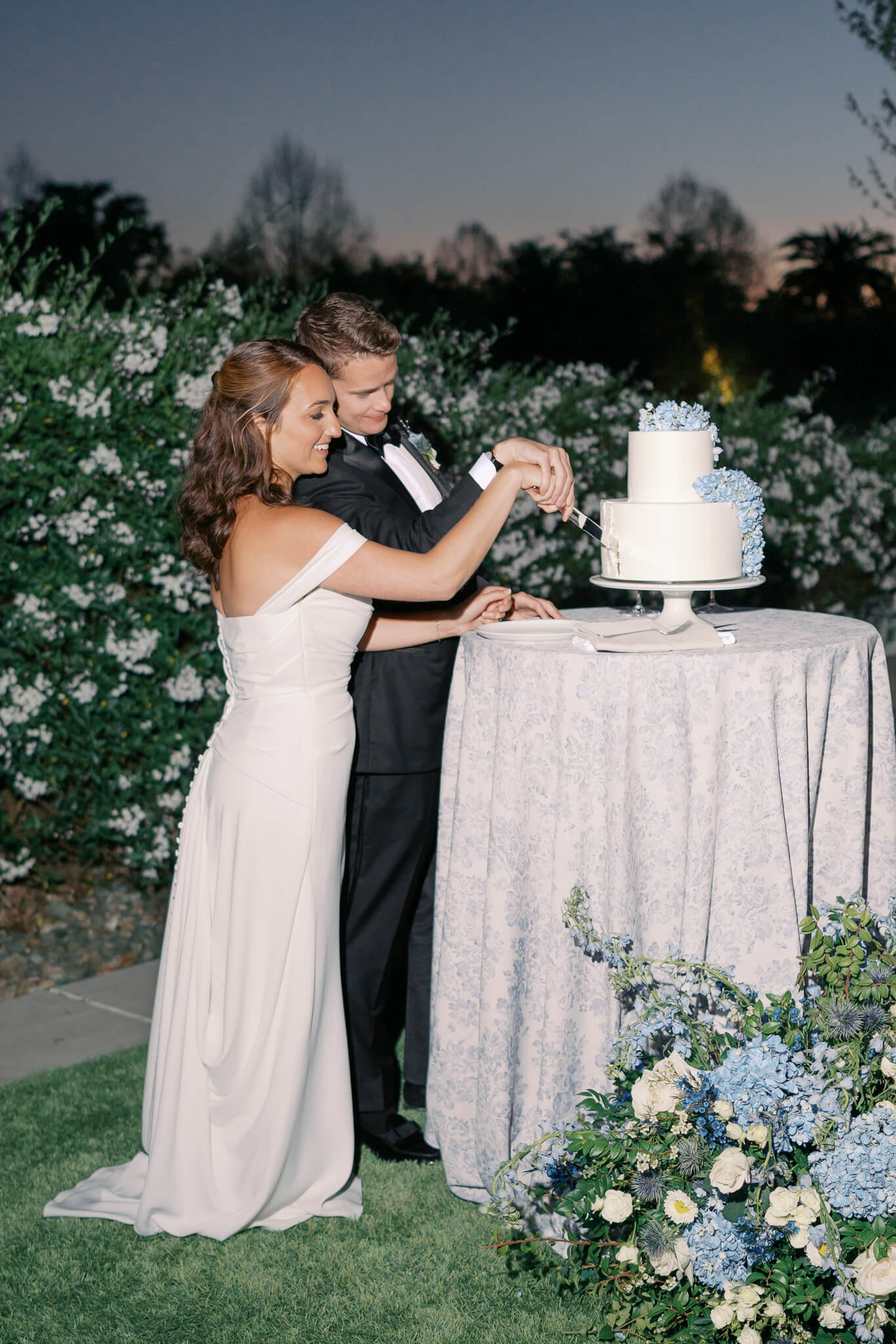 A nighttime shot of a bride and groom cutting their wedding cake surrounded by lush blue and white florals and greenery.