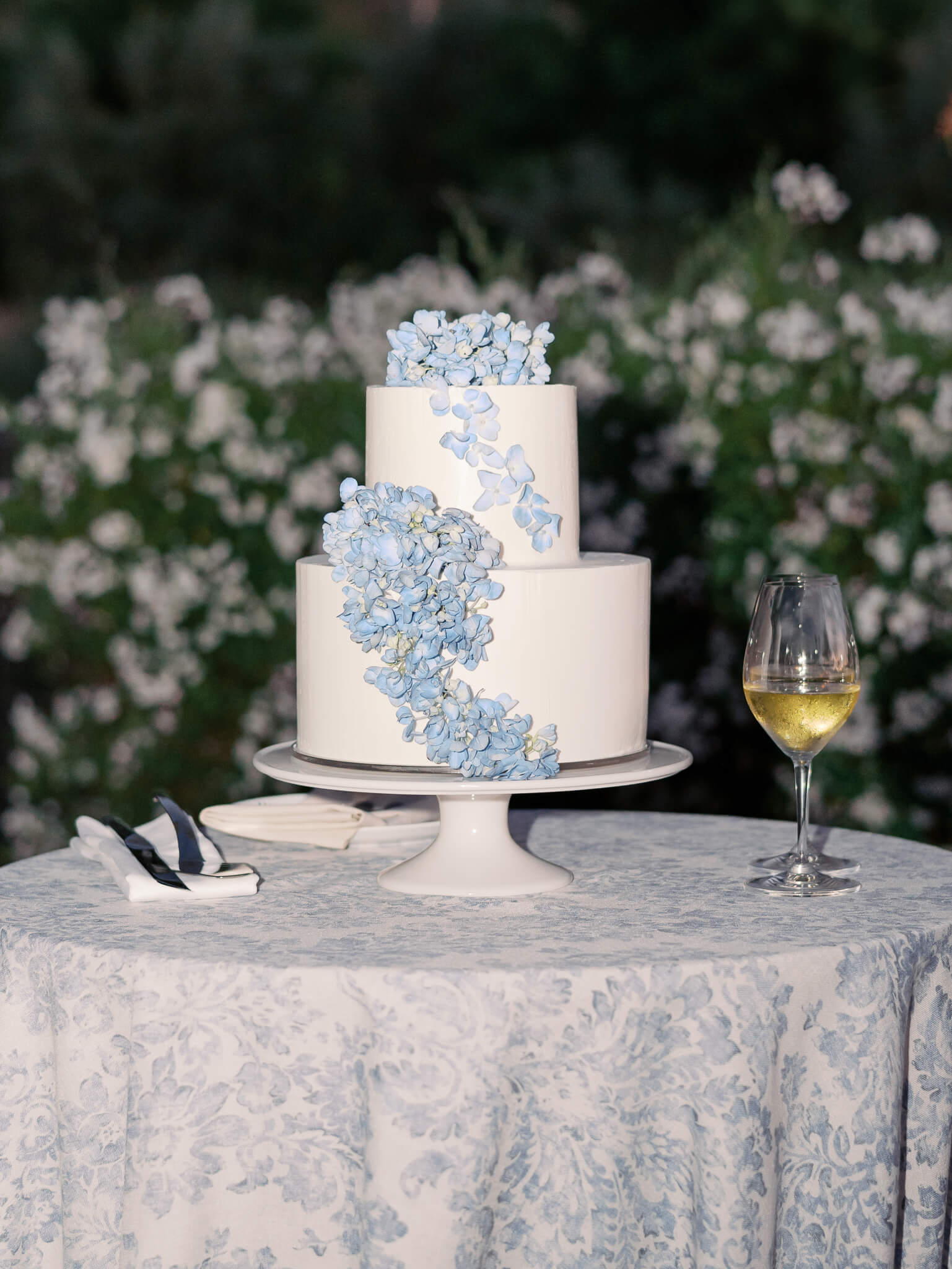 Nighttime image of a white wedding cake with blue hydrangeas sitting on a table with two wine glasses.