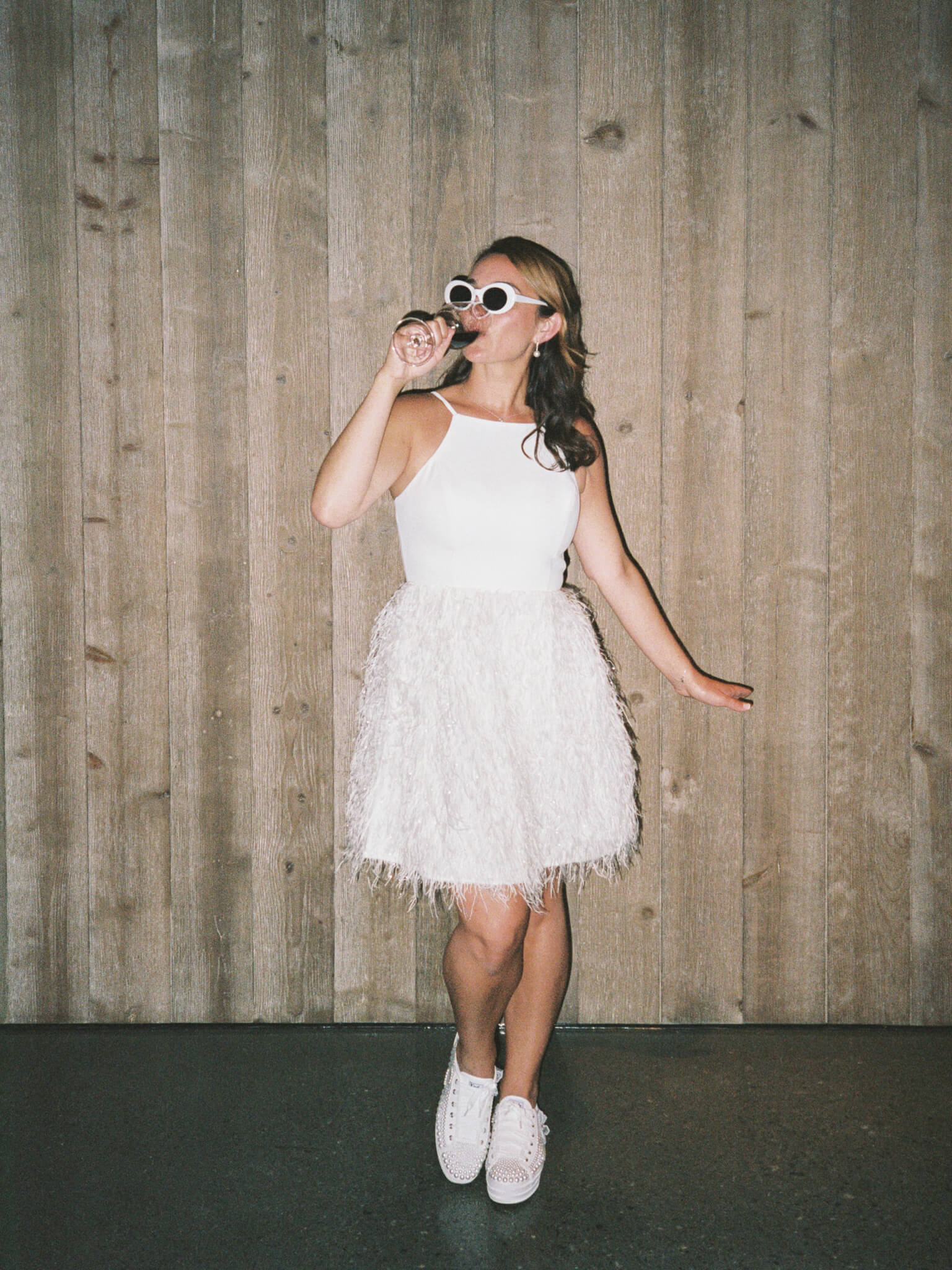 A bride wearing sunglasses and sipping wine standing in front of a wooden backdrop in her short reception wedding dress and sneakers.