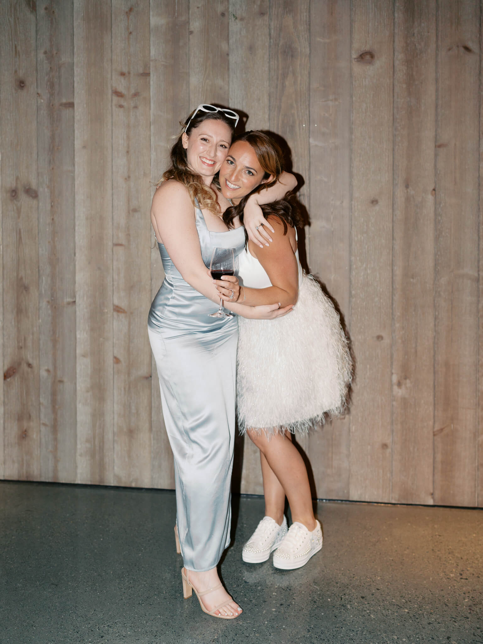 A bride in a short dress and her bridesmaid wearing blue embracing at in front of a wooden backdrop during a wedding reception.