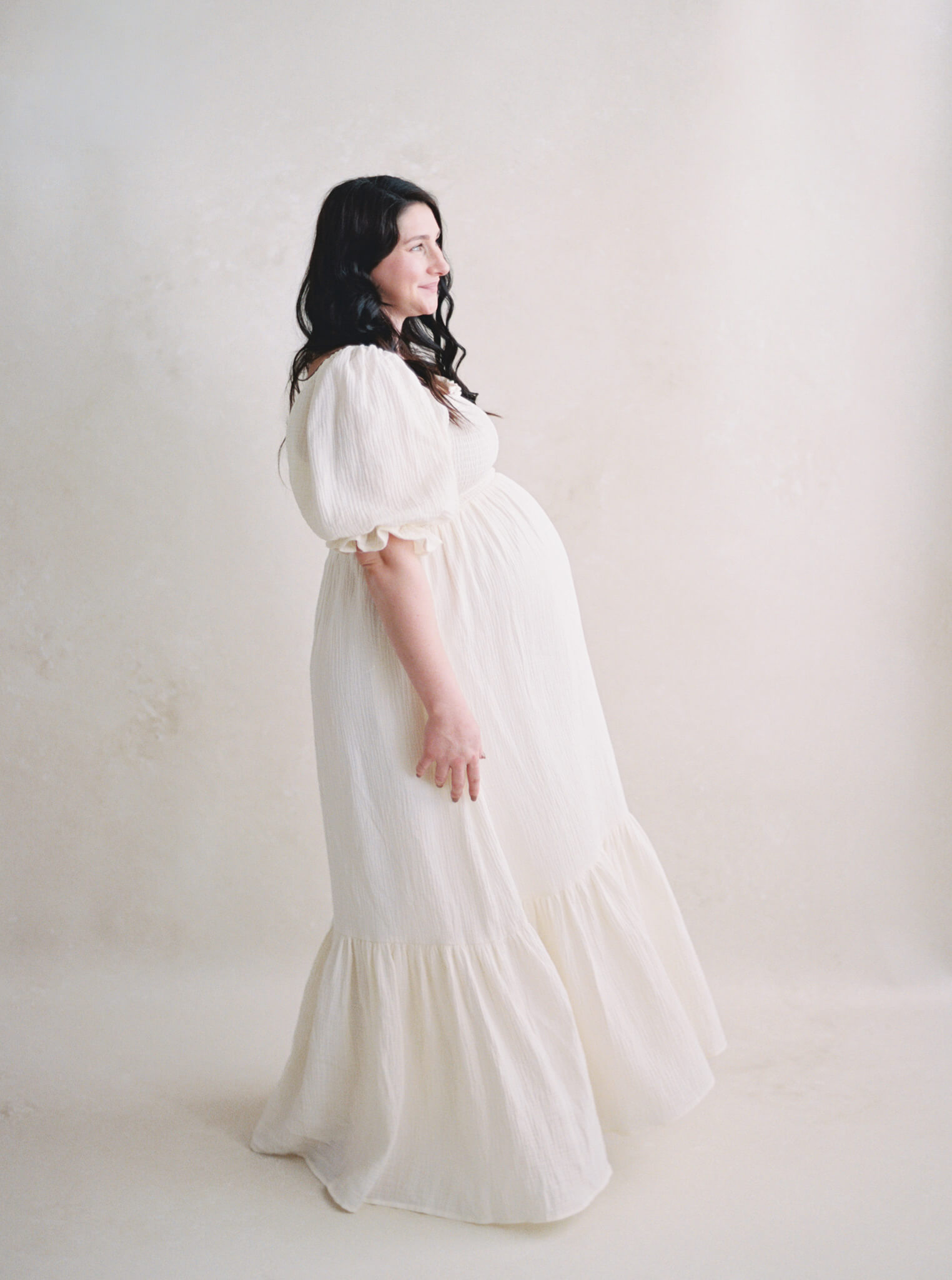 A pregnant woman in a long cream dress turned to the side in front of a neutral backdrop.