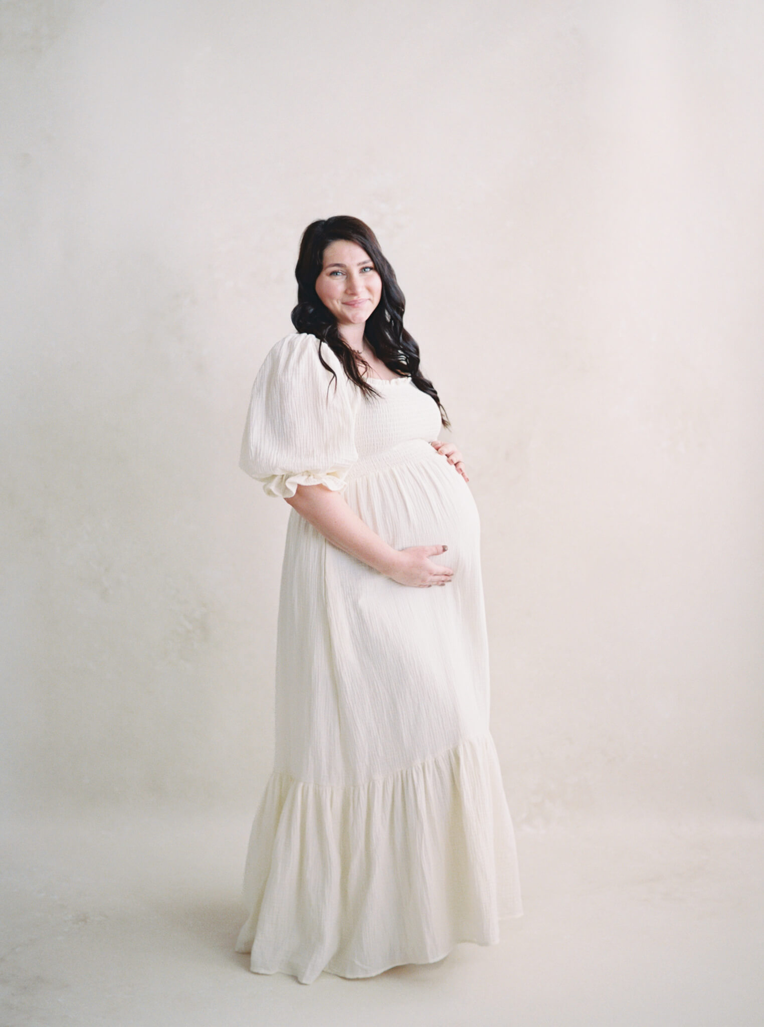 A pregnant woman with dark hair wearing a ling neutral dress and holding her belly.