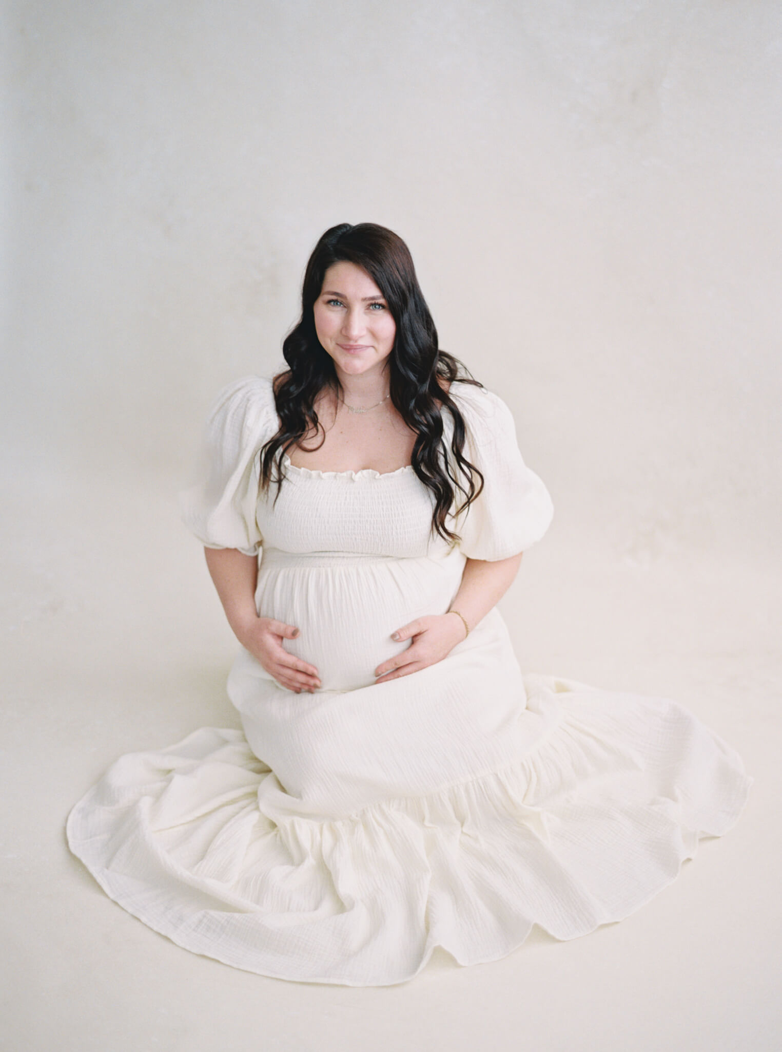 A pregnant woman with dark hair and a long cream dress sitting in front of a neutral backdrop.