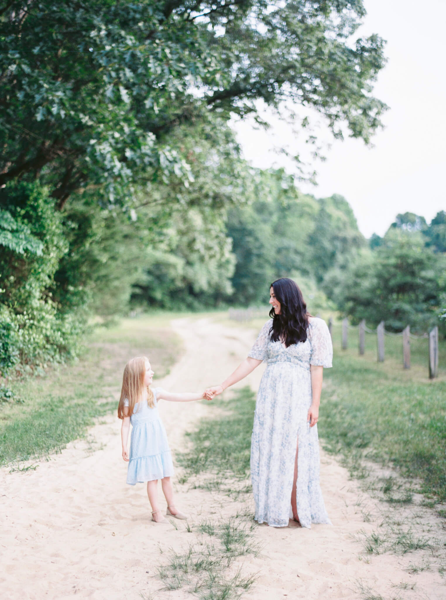 A mom and daughter in pale blue dresses holding hands looking at each other while standing on a sandy path under an overhanging tree during their Annapolis Family Photos.