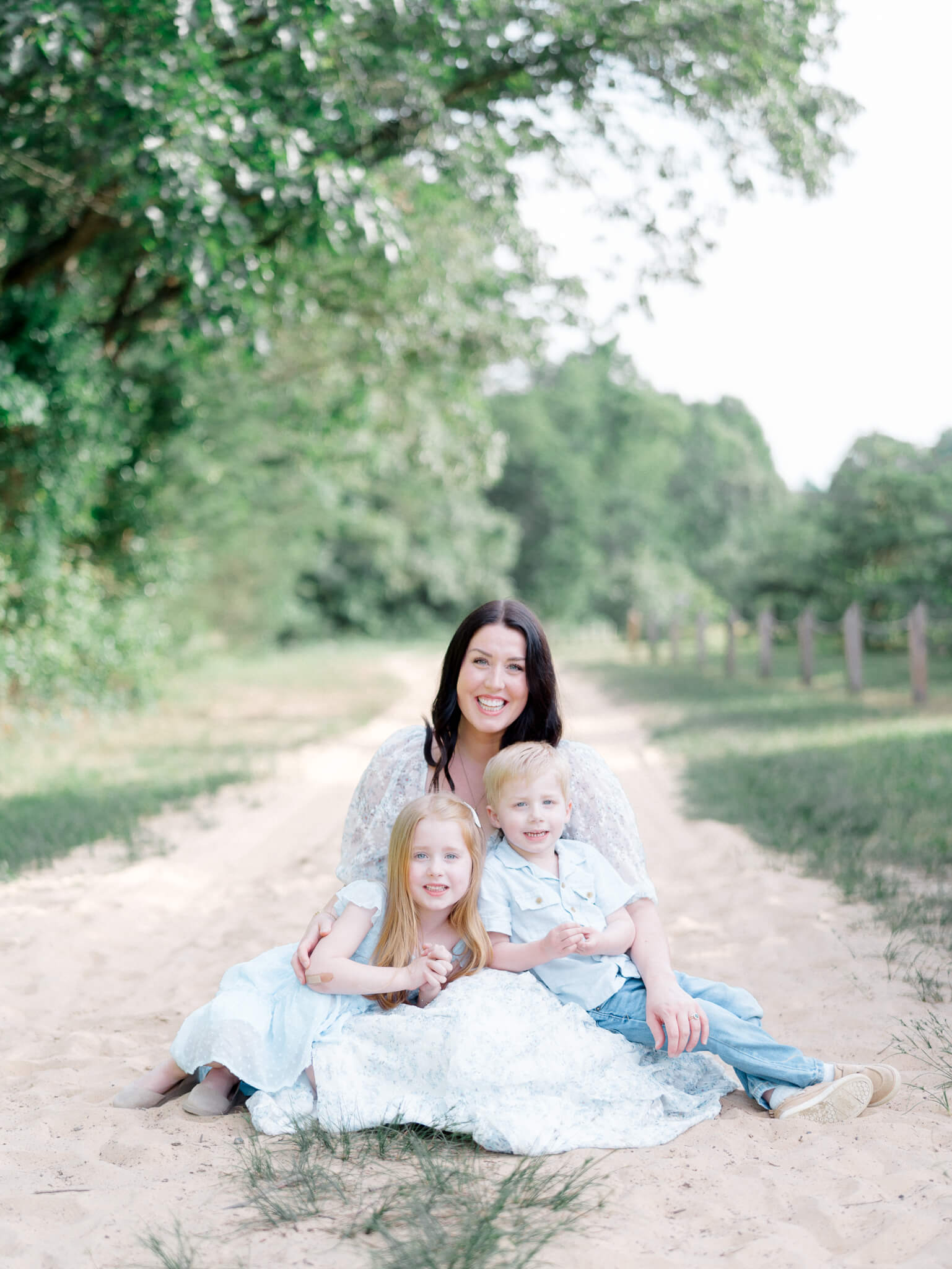 A mother in a blue dress sitting on a sandy path with her son and daughter on her lap during their Annapolis Family Photos.