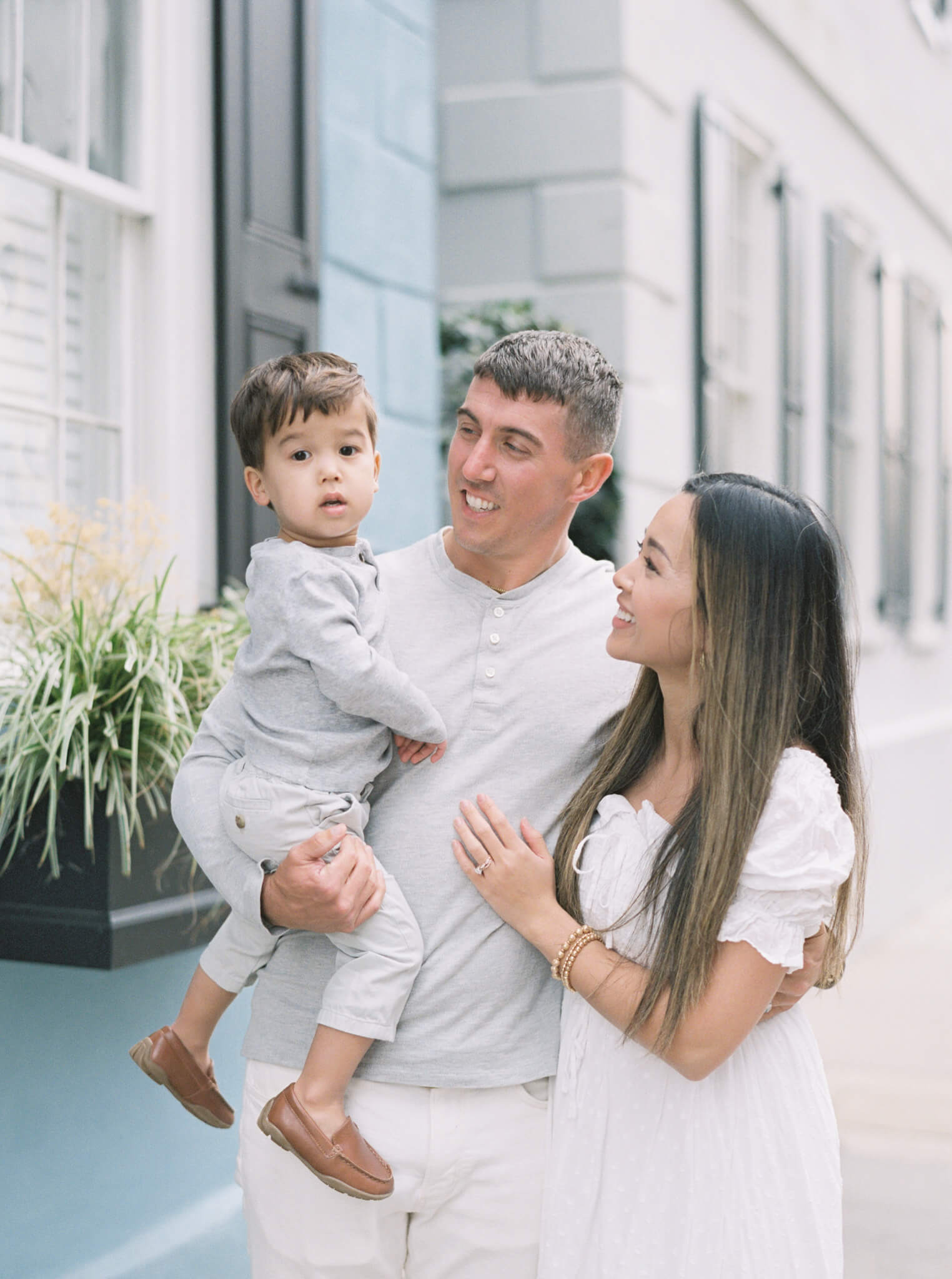 A mom in a white dress and dad in a gray shirt and khakis holding their son and smiling at him, who is looking at the camer.