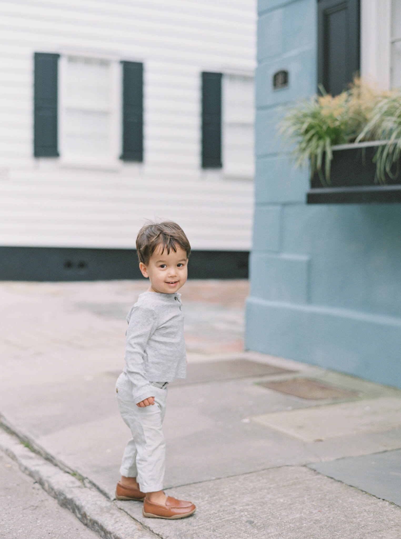 A little boy standing on a sidewalk during a Charleston Family Session turns and smiles at the camera.