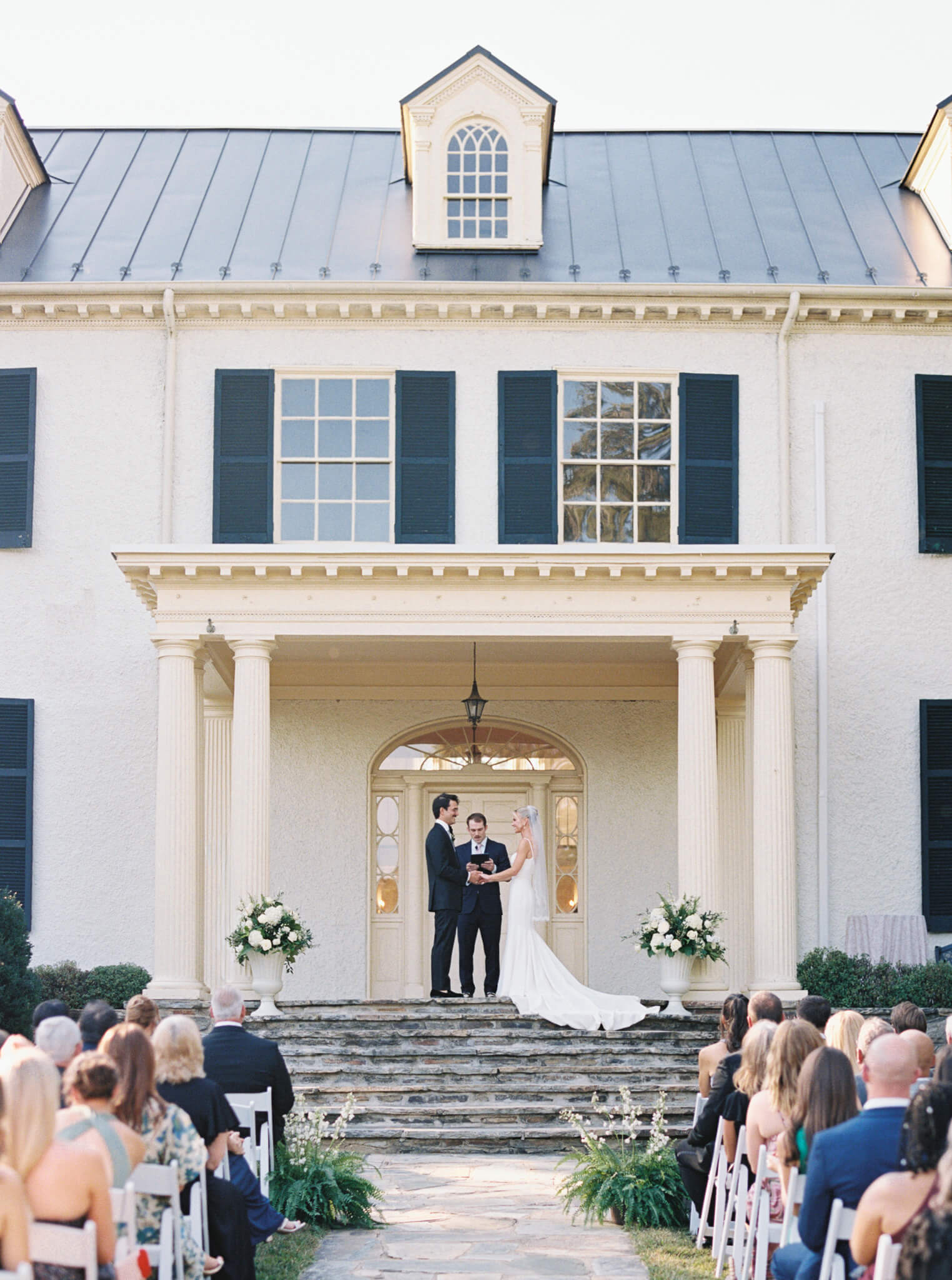 A bride and groom holding hands during their ceremony on the back patio of Rust Manor House while their guests are watching.