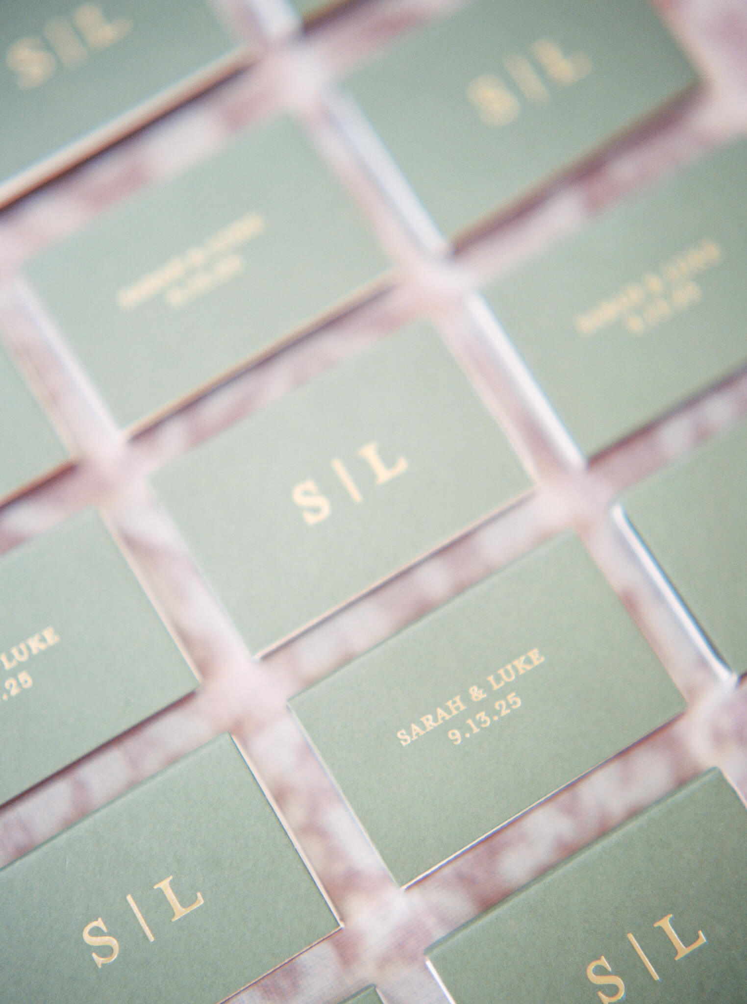 Wedding flatlay image of sage green matchboxes arranged in rows with gold lettering of the couple's names, initials, and wedding date on a pink background.