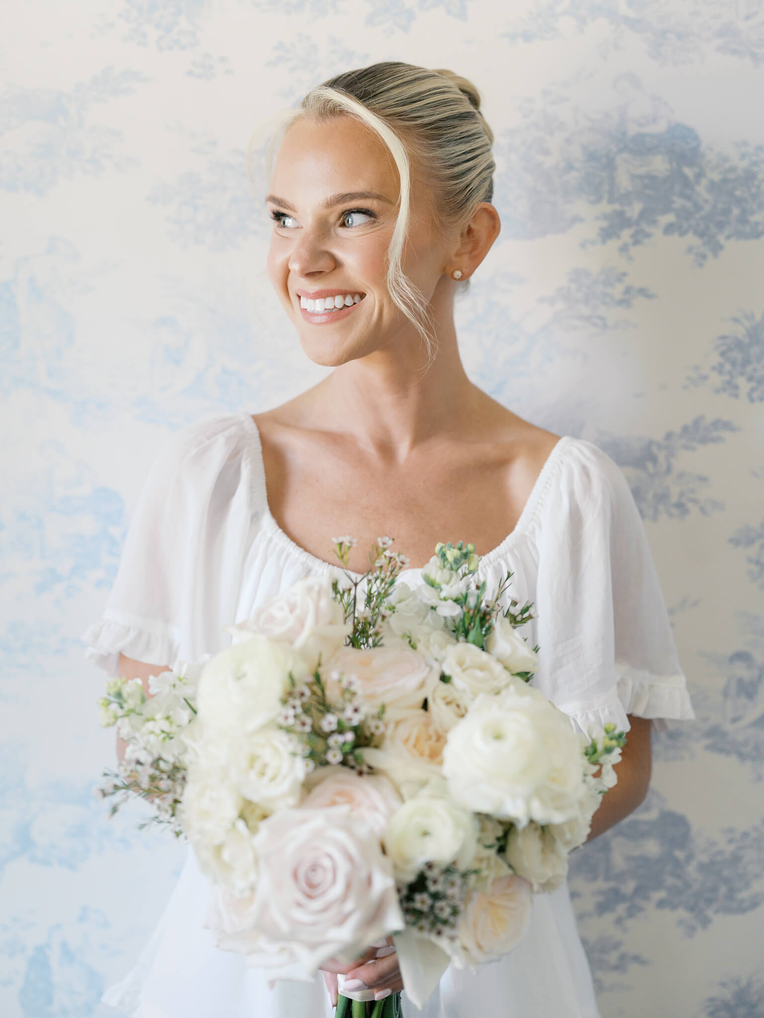 Closeup image of a bride wearing white linen pajamas and holding her bouquet while smiling to the side in front of a blue and white toile background at her Rust Manor House wedding.