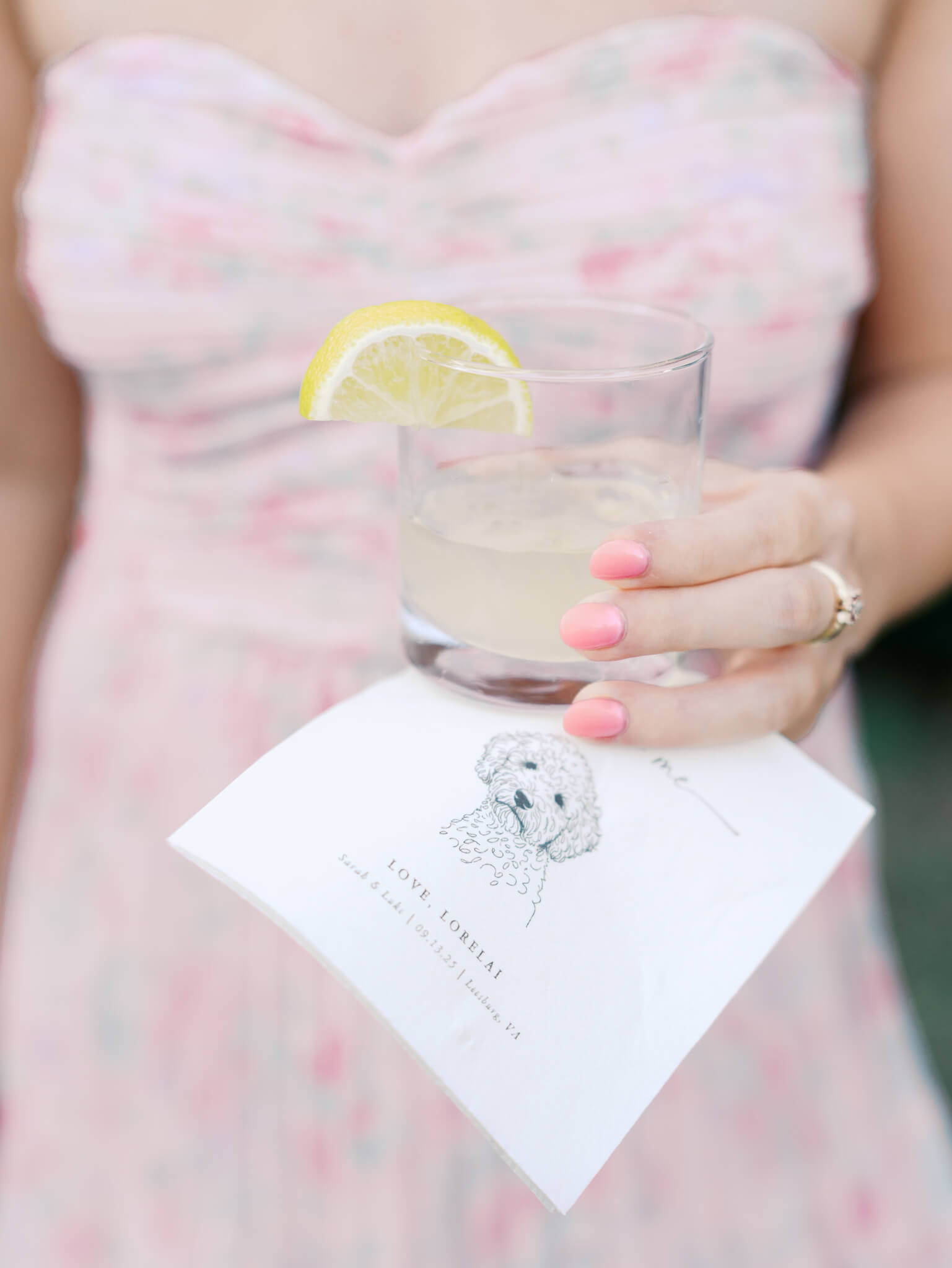 Closeup image of a wedding guest's hand wearing pastel pink holding a yellow drink with a lemon and cocktail napkin featuring a dog.