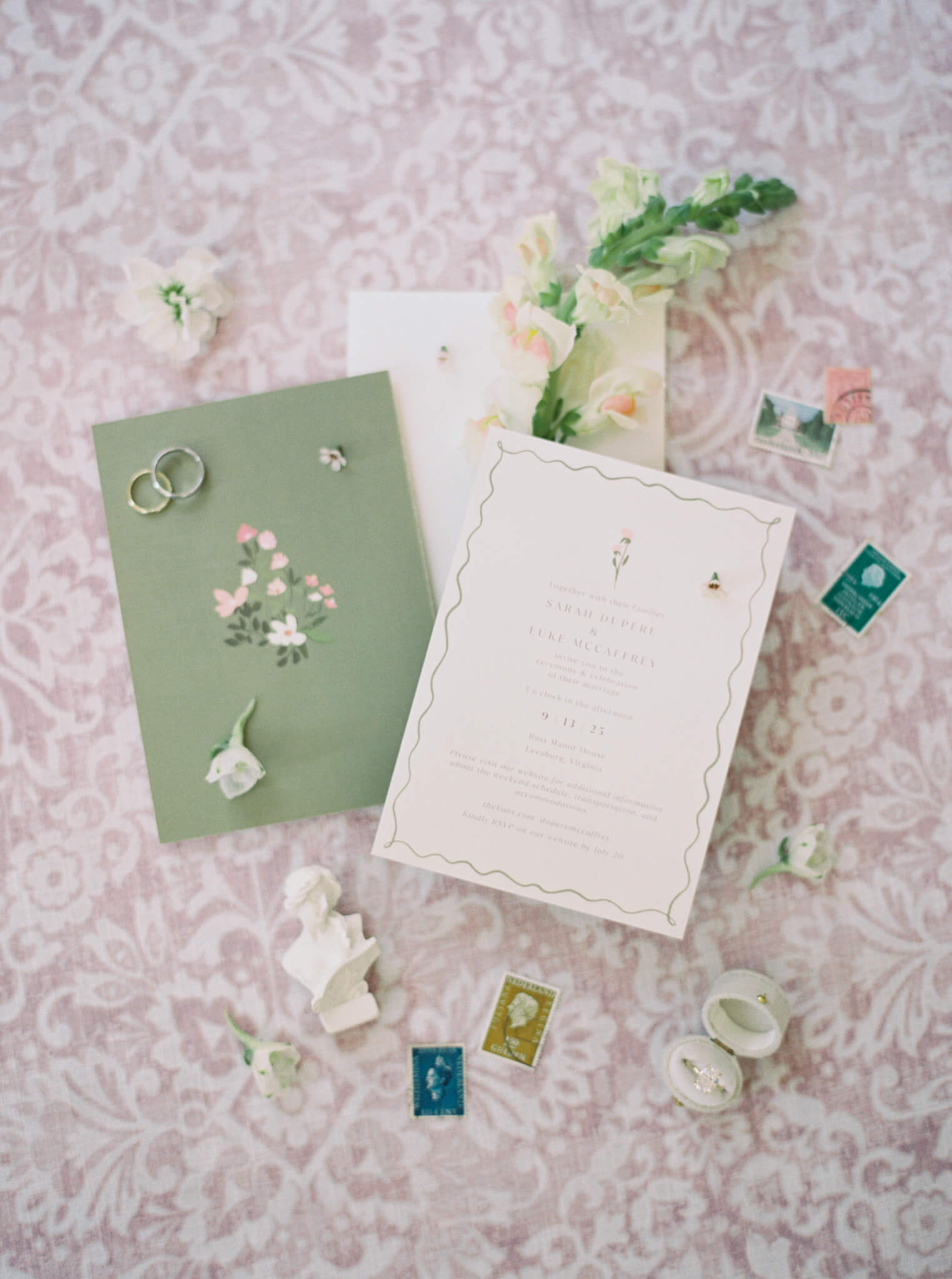 Flatlay image of a wedding invitation suite in greens, pinks, and creams with stamps, rings, and flowers on a pink and cream damask background.